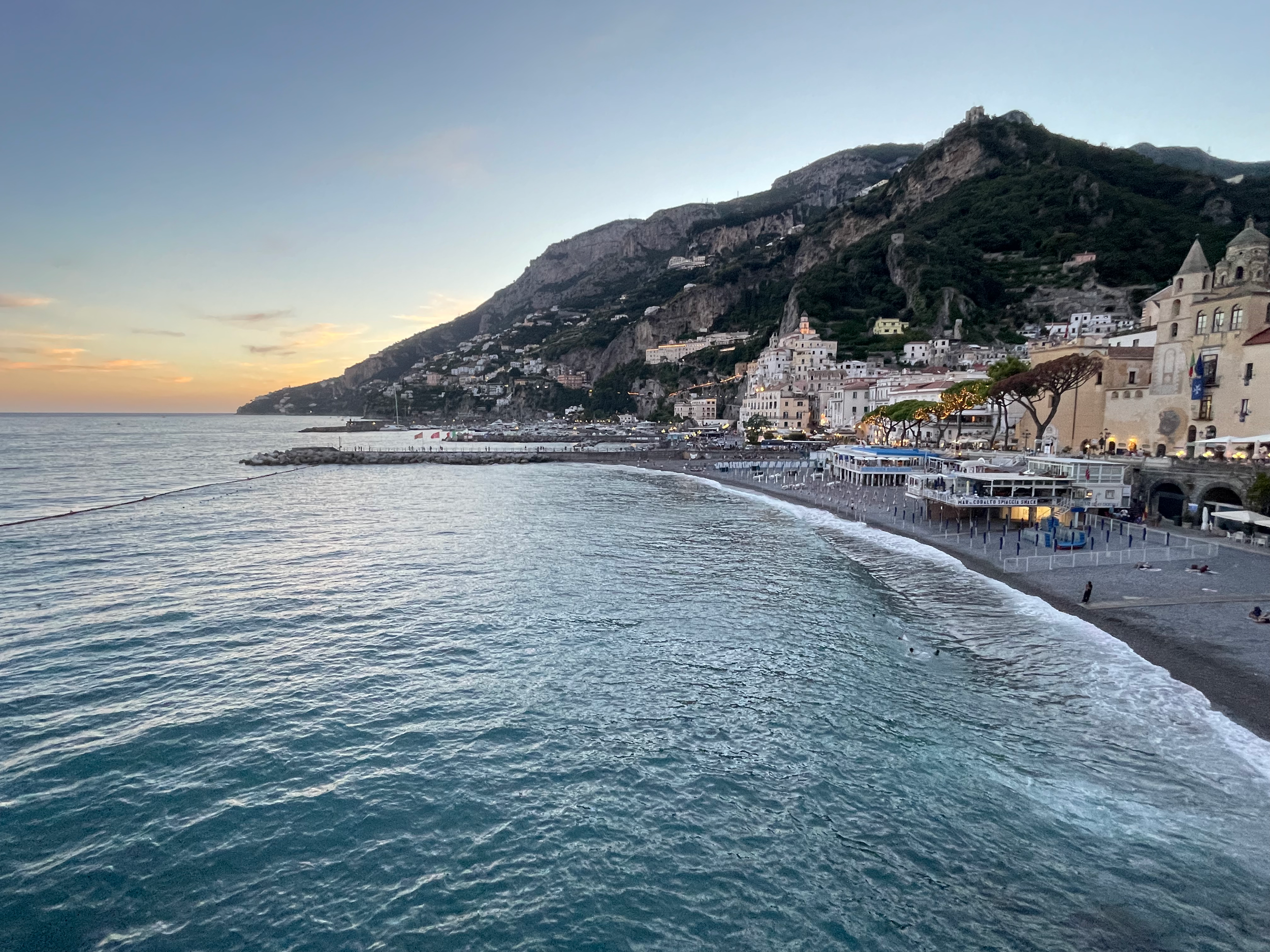 Scenic view of a coastal town built on a hillside with white buildings, a church with a bell tower, and a promenade along the water at sunset.