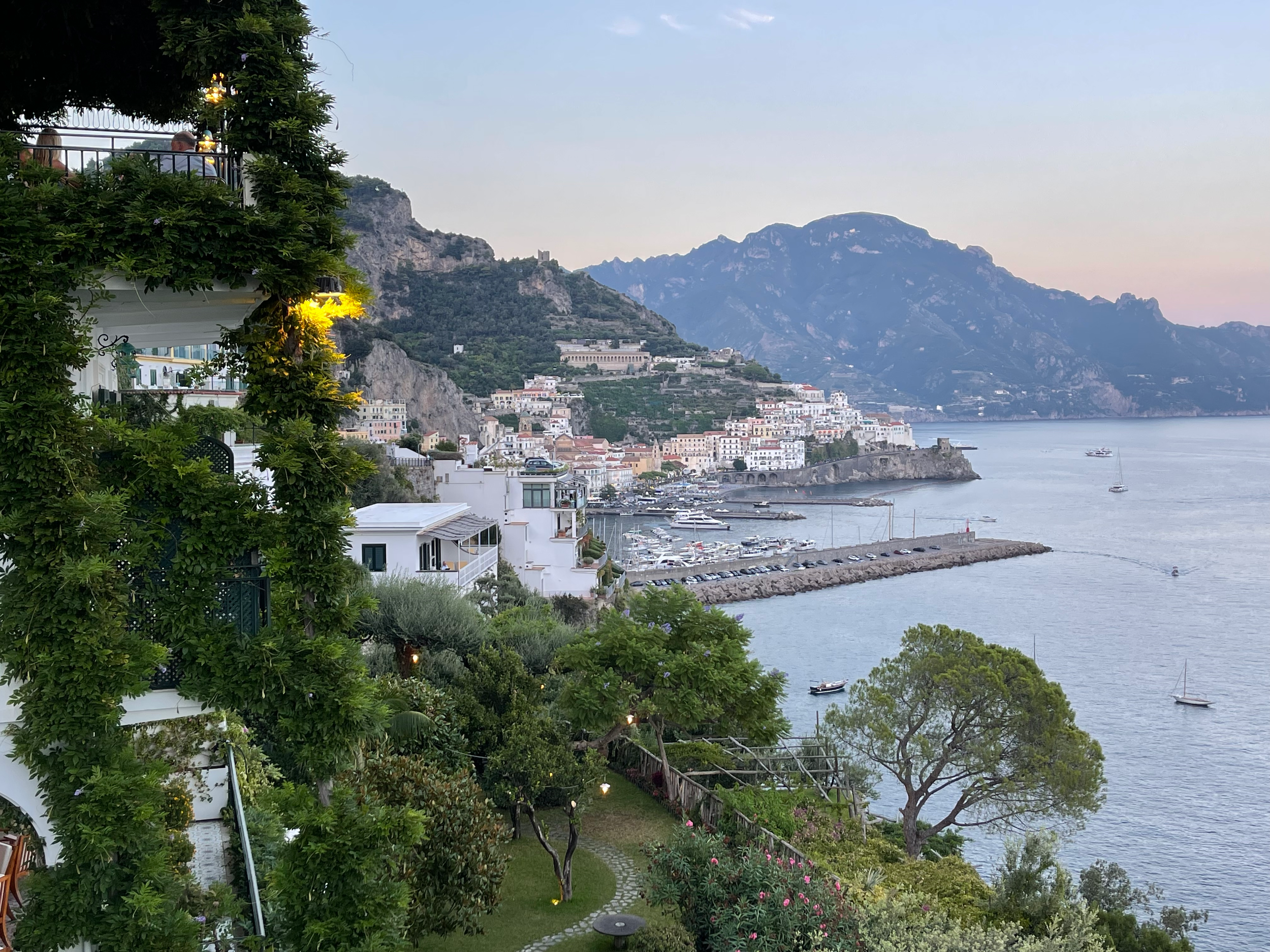 A scenic view of a coastal town with white buildings on a hillside, a calm bay with boats, and mountains in the background, seen from a balcony with greenery and outdoor lighting.