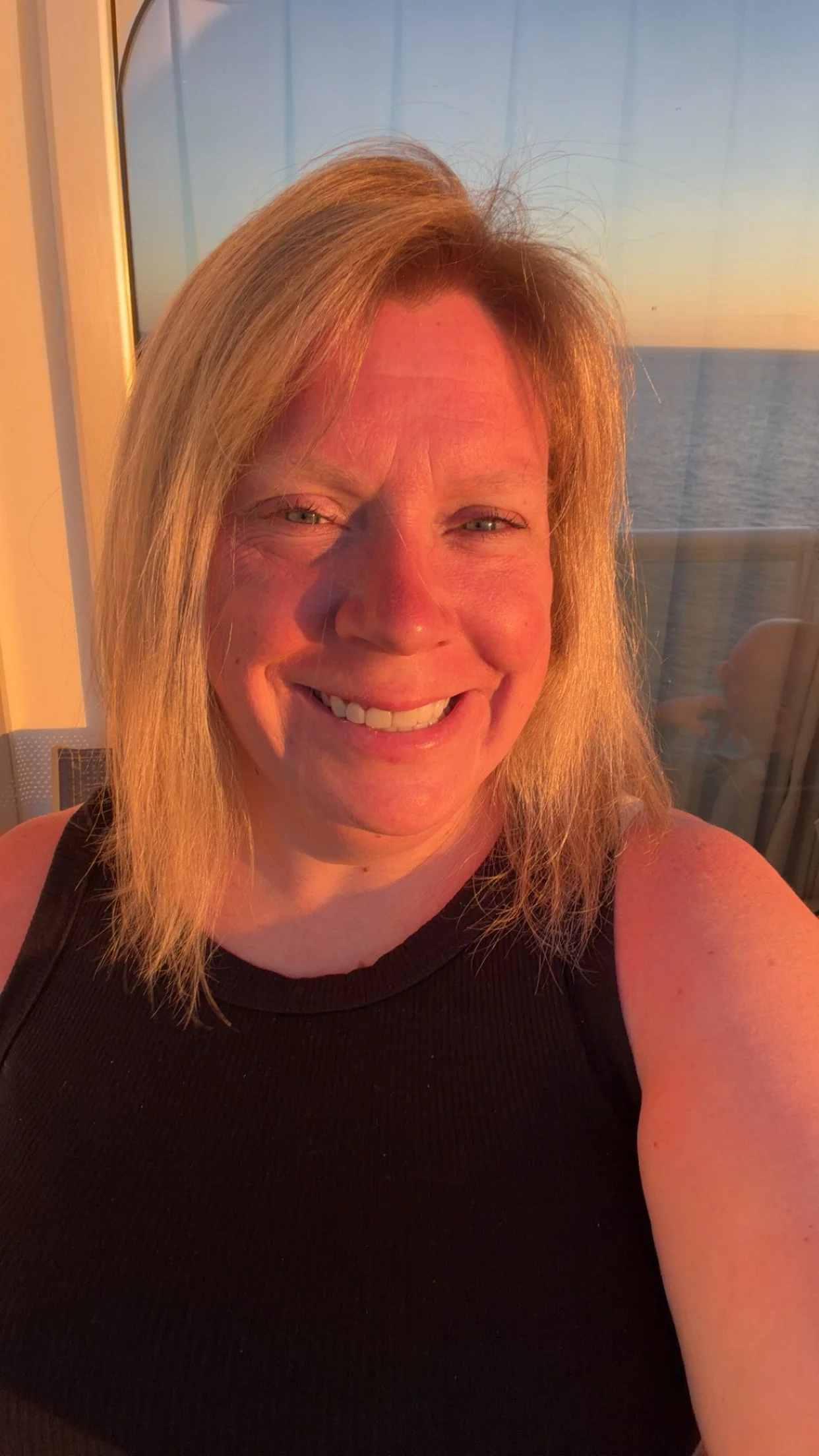 A smiling woman with blonde hair takes a selfie on a balcony at sunset, with a view of the ocean in the background.