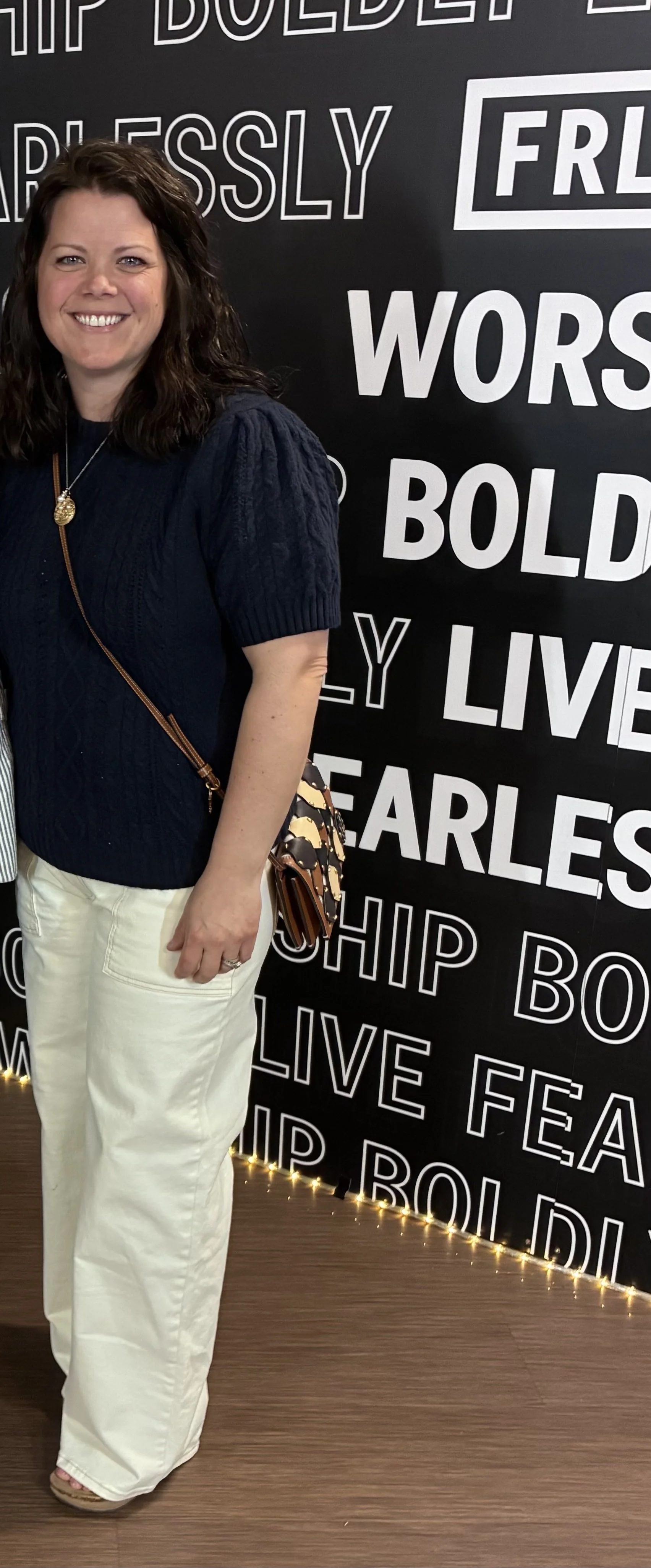 A woman with curly brown hair smiling, wearing a navy blue sweater, white pants, and a necklace, standing next to a black wall with white text.