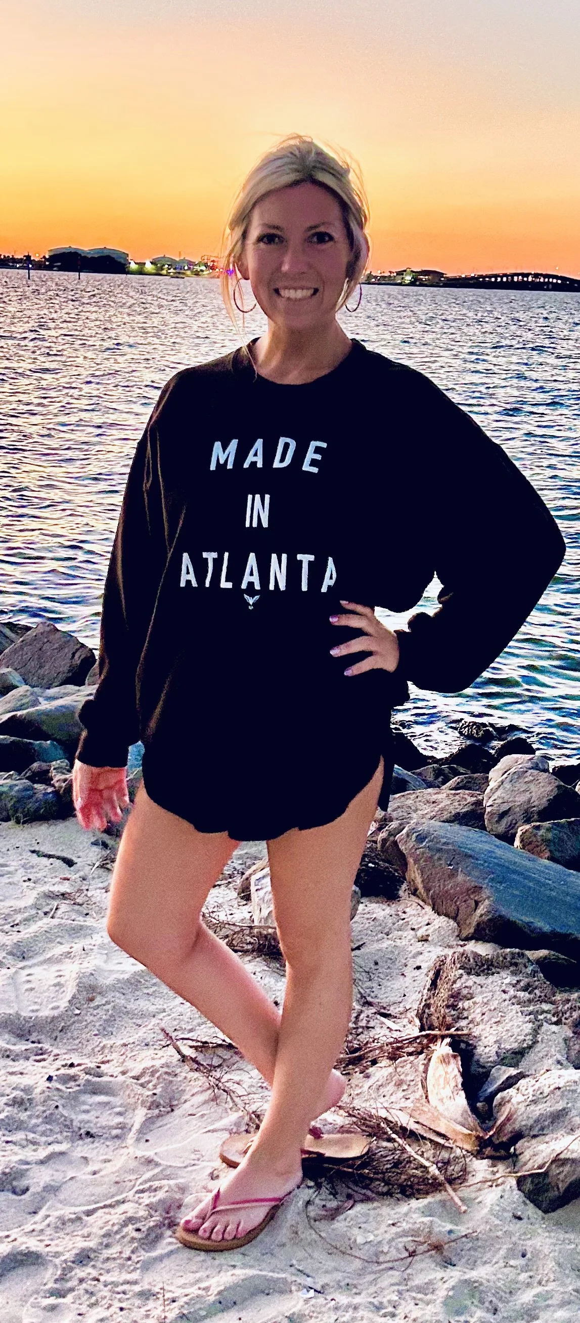 A woman standing on a rocky beach at sunset, wearing a black sweatshirt that says 'Made in Atlanta', black shorts, and pink flip-flops, with her hand on her hip and smiling.