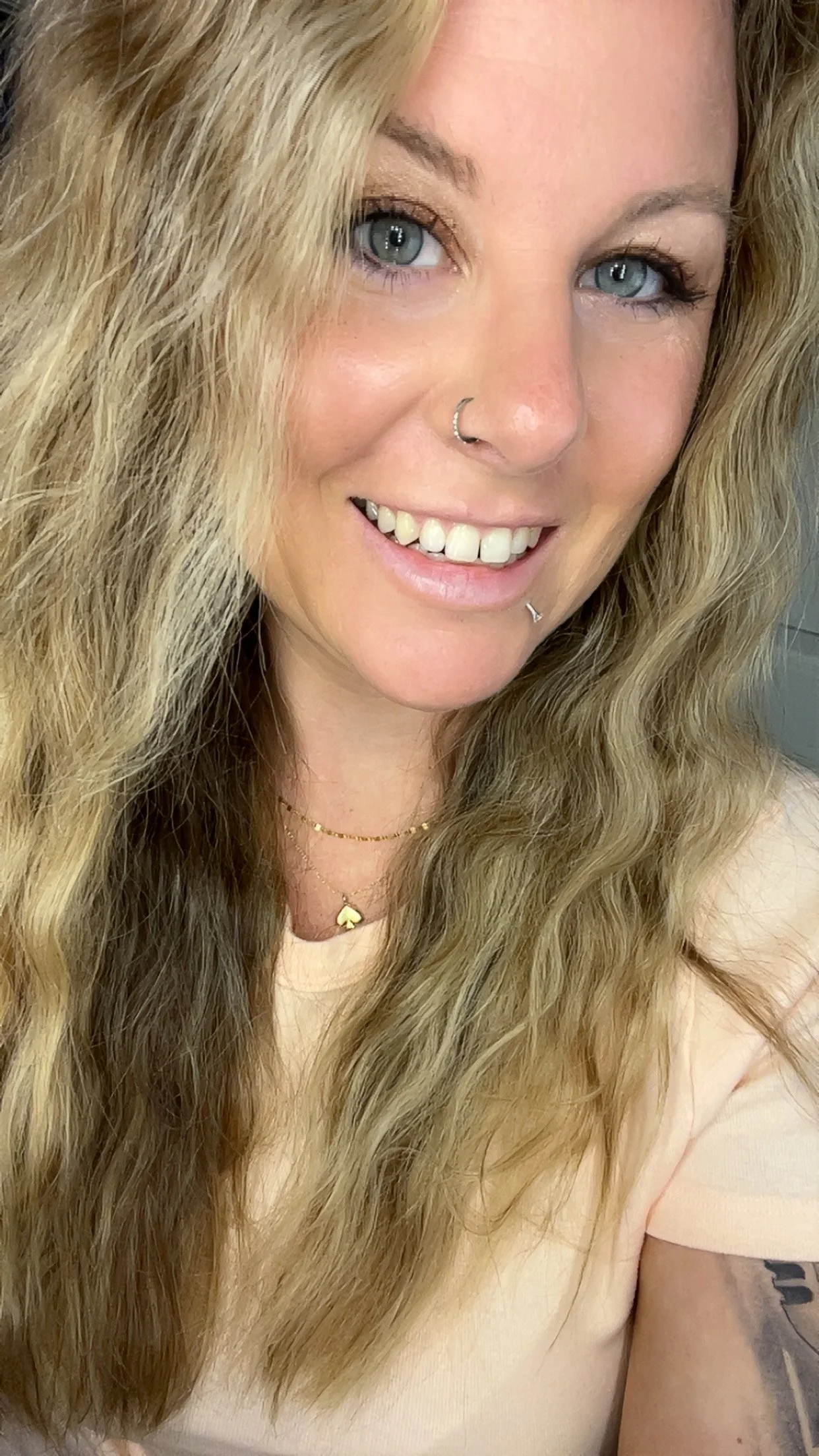 A woman with long, wavy blonde hair, blue eyes, and a nose and lip piercings, smiling at the camera, wearing a beige top and gold jewelry.