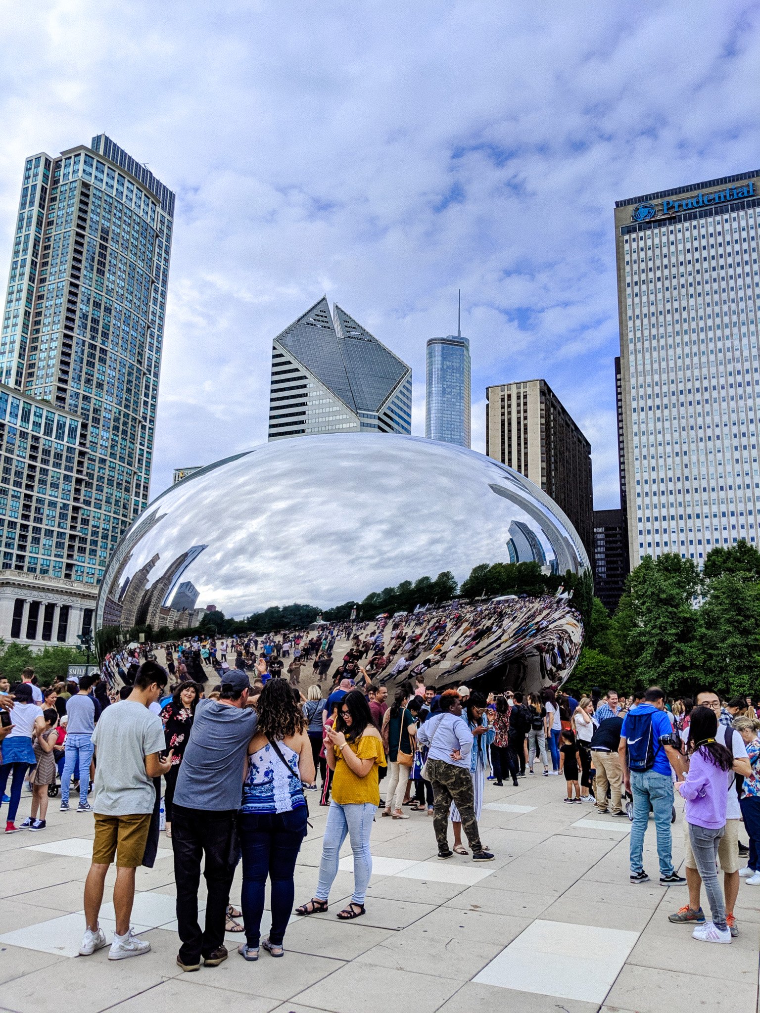 A crowd of people gathered in a park near the Cloud Gate sculpture in Chicago, surrounded by tall city buildings, with partly cloudy skies overhead.