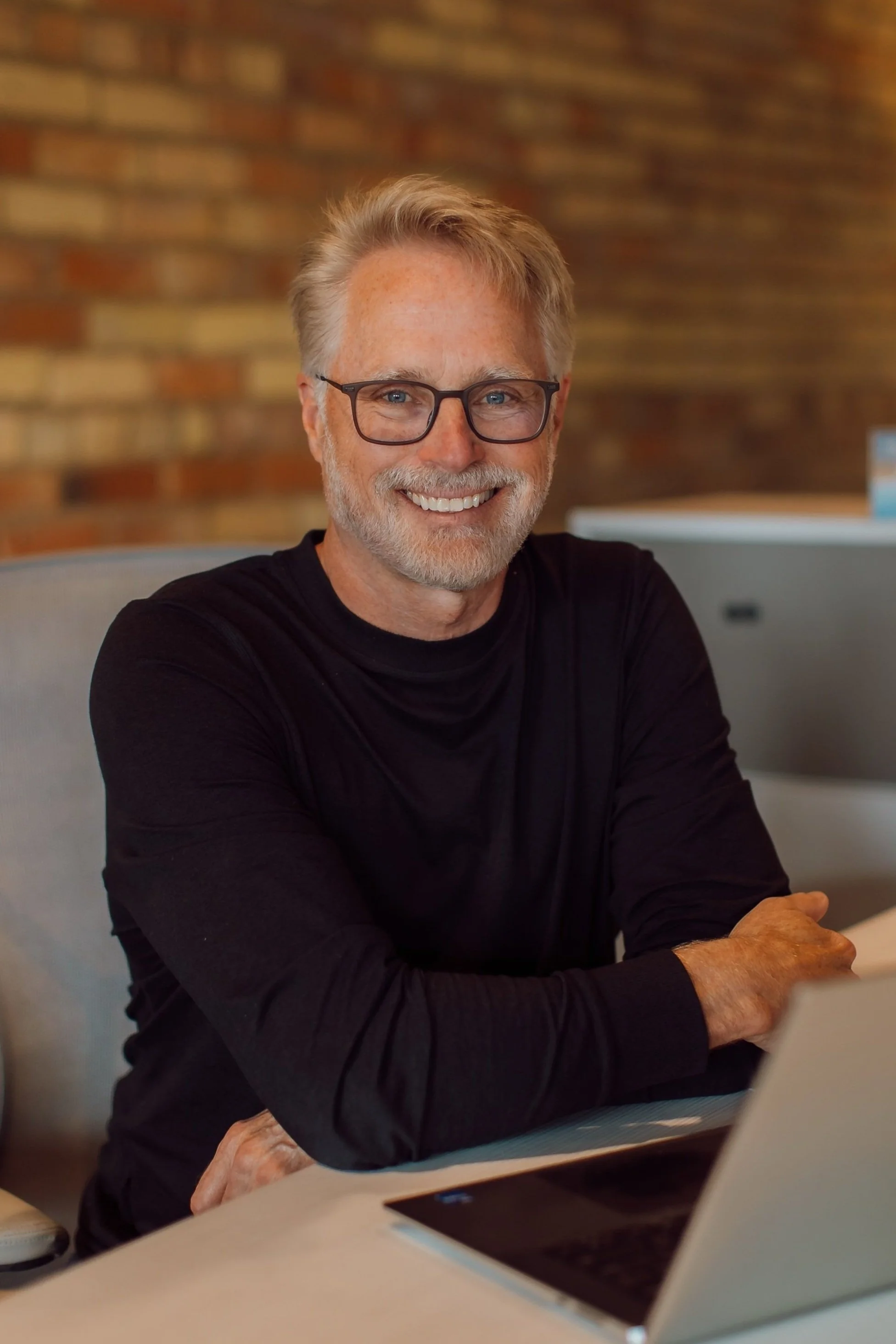 A smiling middle-aged man with gray hair and beard, wearing glasses and a black shirt, sitting at a table with a laptop in a cozy indoor setting with a brick wall background.