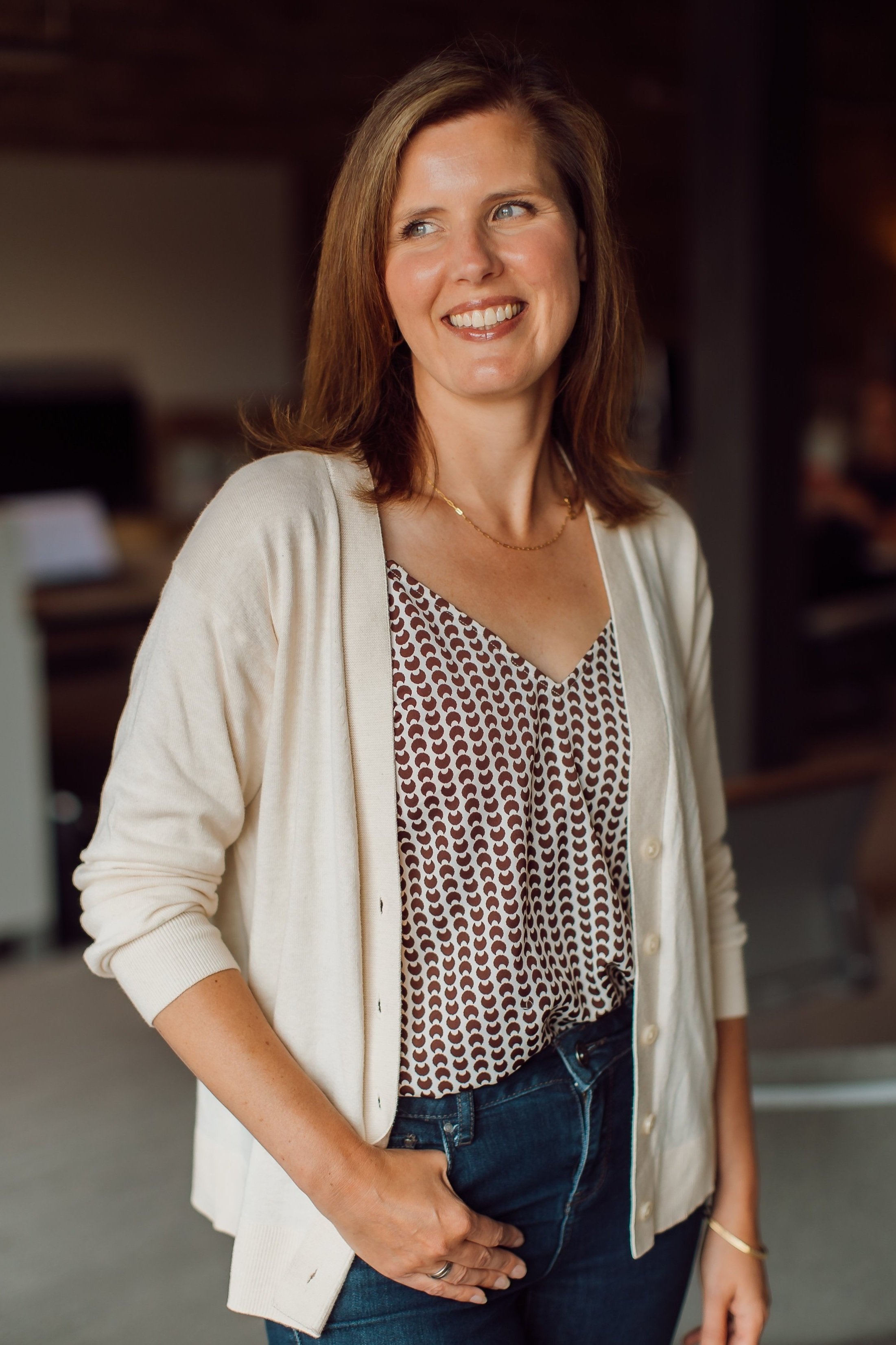 A woman with brown hair wearing a patterned blouse, cream cardigan, and jeans, smiling and standing indoors.