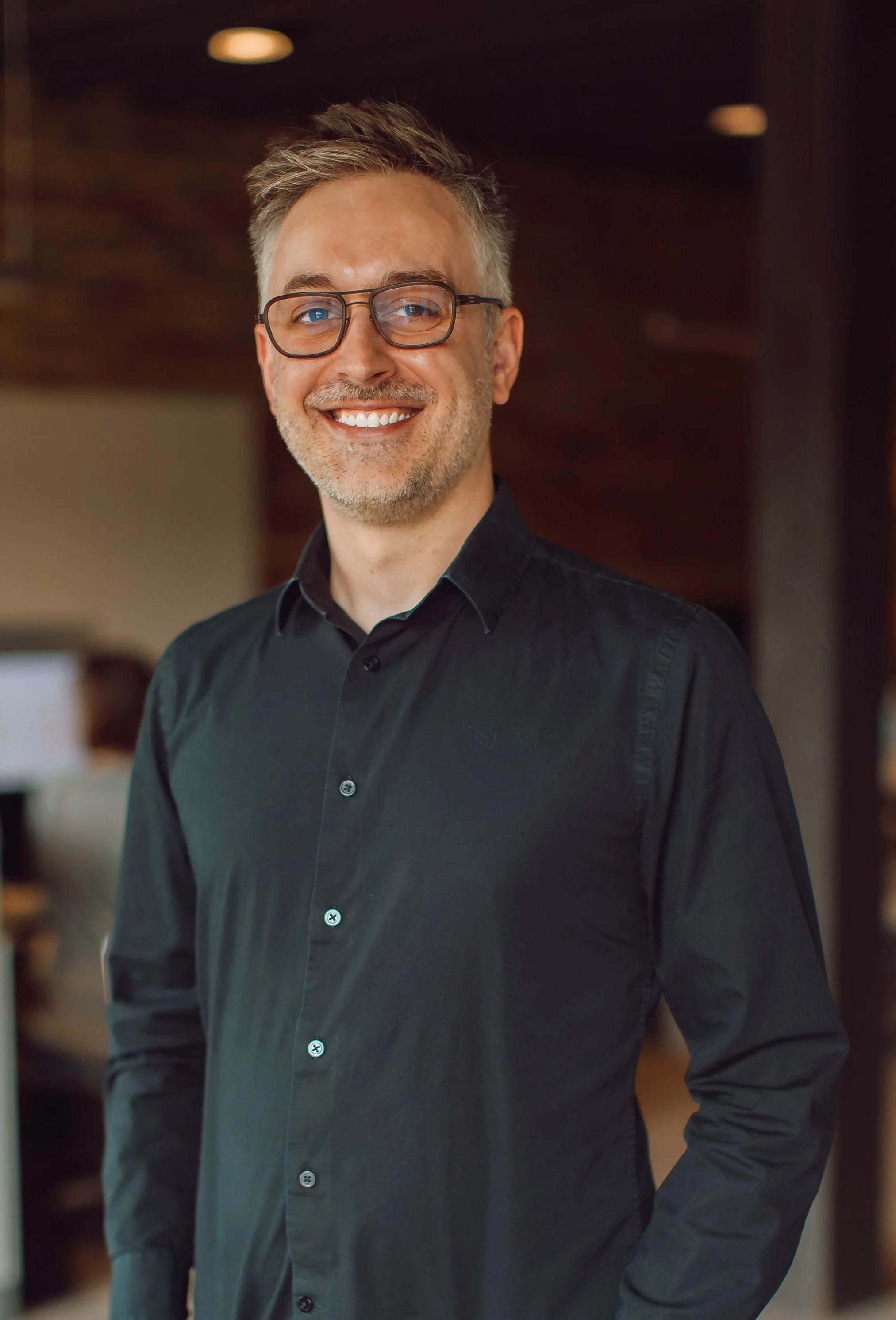 A portrait of a smiling middle-aged man with light-colored hair, beard, and glasses, wearing a black shirt, standing indoors with a blurred background.