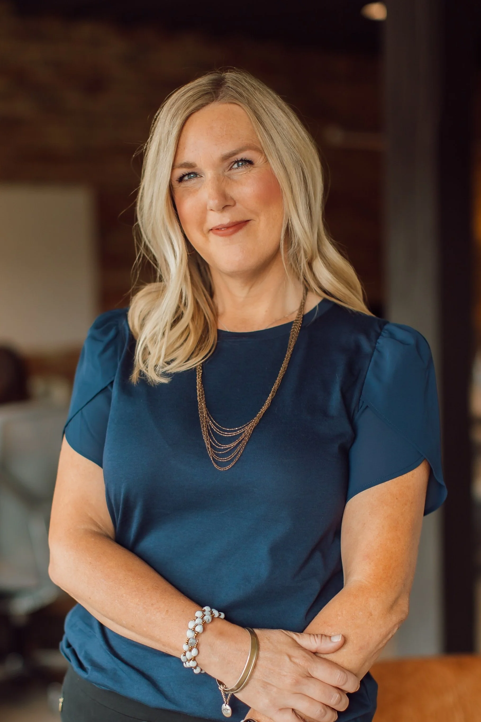 A woman with blonde hair wearing a dark blue top, layered necklaces, and bracelets, smiling while standing with crossed arms in a warmly lit indoor setting.