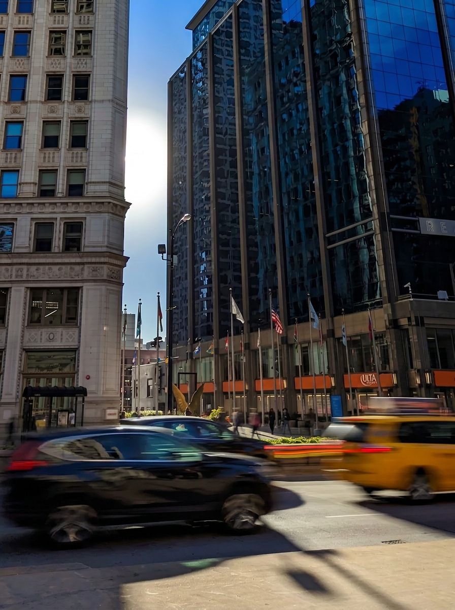 City street scene with tall modern glass buildings, flags on poles, blurred moving cars including a black vehicle and a yellow taxi, and pedestrians walking on the sidewalk. The sky is clear and the sun is reflecting off the buildings.