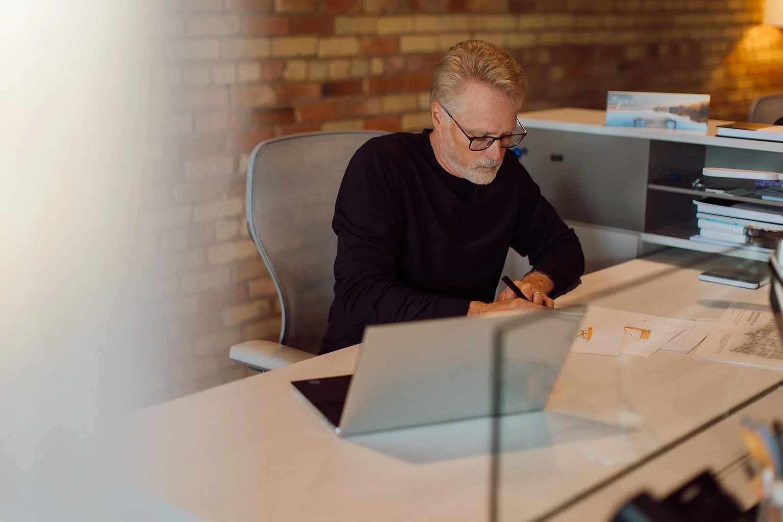 An older man with glasses and gray hair sits at a desk in a modern office, working on papers with a pen. There are books and a laptop on the desk with a brick wall background.