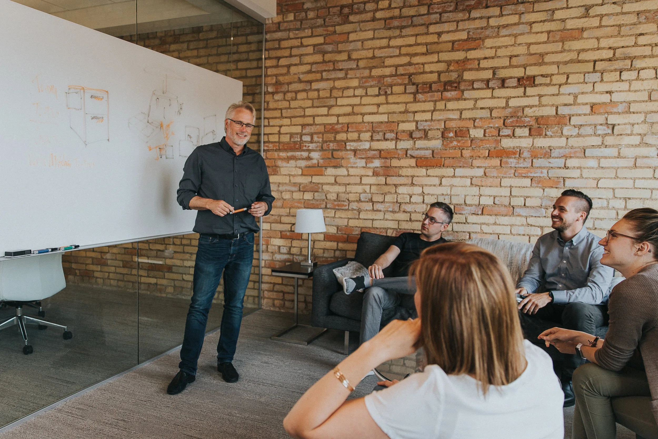 A man in a black shirt and glasses is standing in front of a whiteboard and presenting to a group of people sitting on a sofa and chairs in a room with a brick wall. The group is engaged and smiling.
