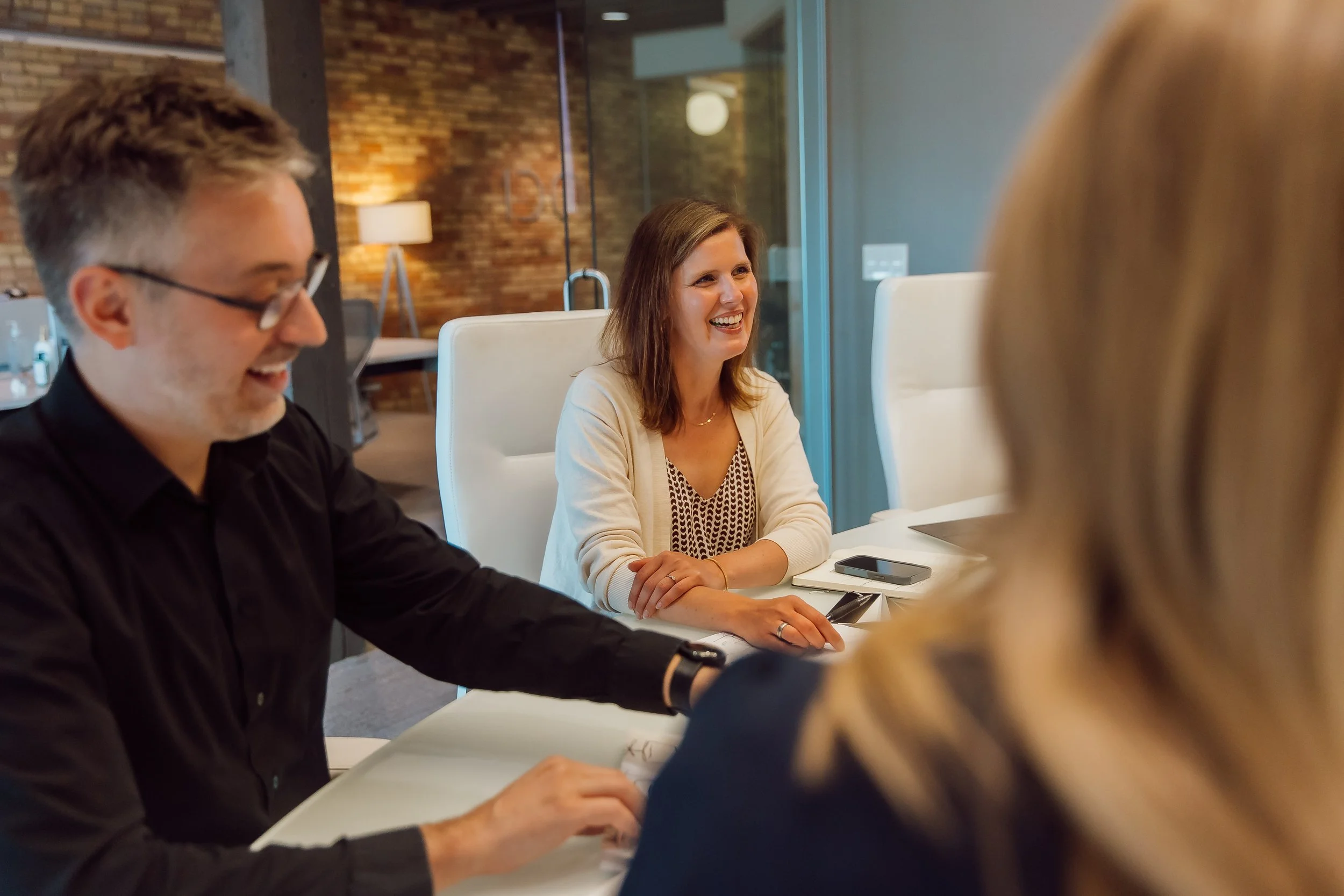 Group of people sitting around a white conference table in a modern office, engaged in a discussion, smiling, with a woman in the center talking, a man on the left and a woman with blonde hair in the foreground.