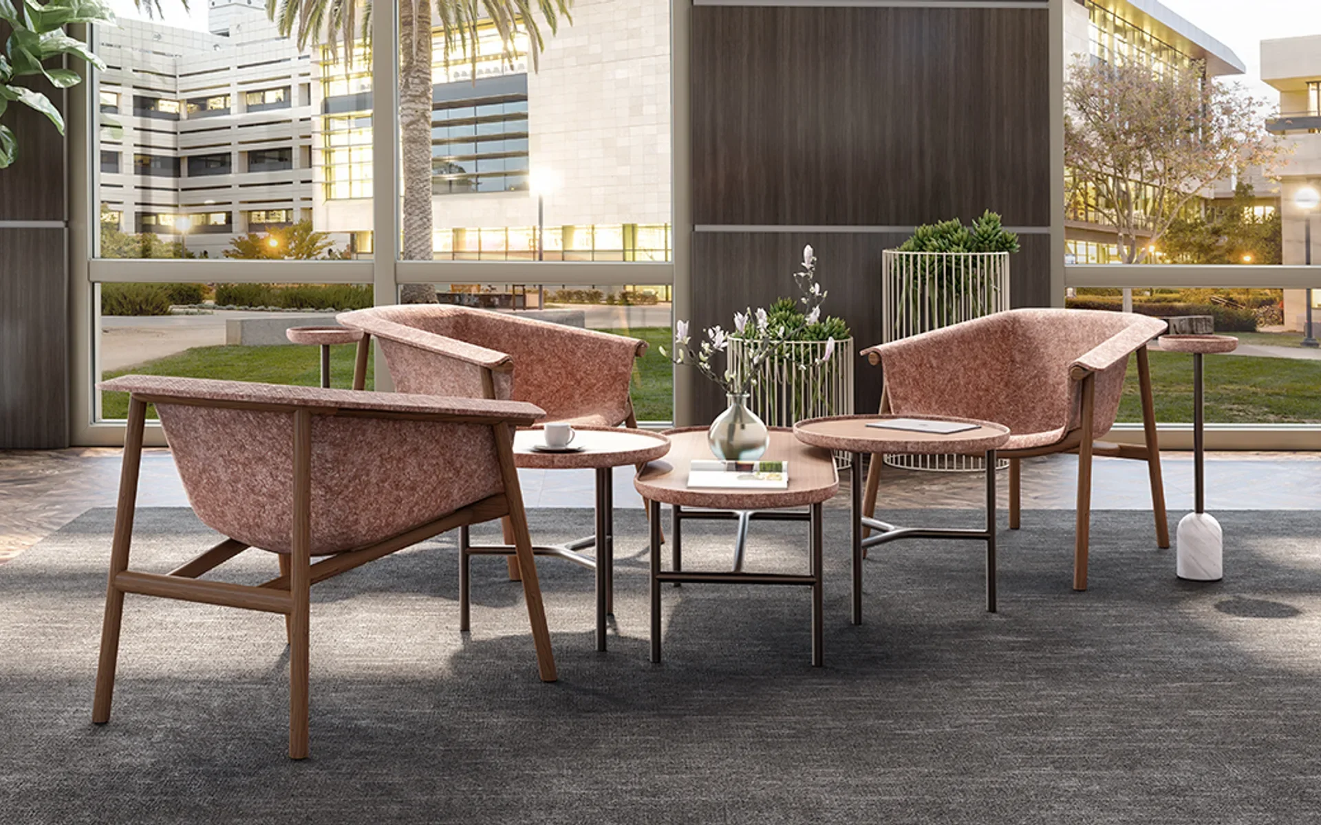 Modern lounge area with pink upholstered armchairs, small table with a vase of white flowers, and large windows showing an outdoor plaza with trees and a building.