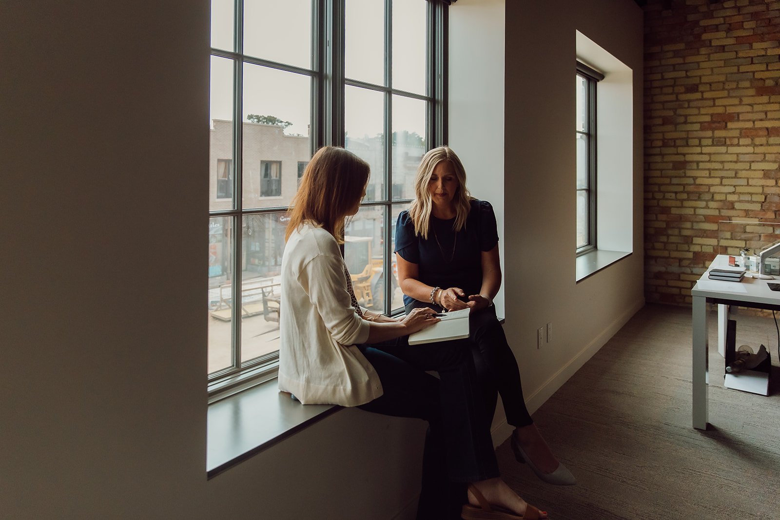 Two women sitting by large windows, engaging in conversation or a meeting, with a city street visible outside.