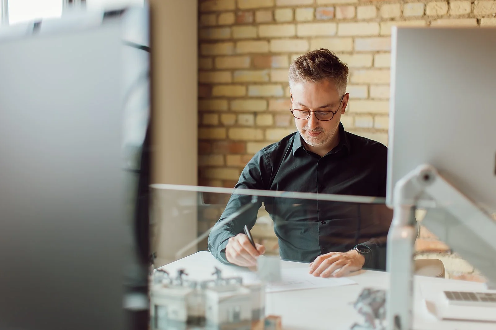 A man with glasses and a black shirt working at his desk, writing with a pen. The scene is viewed through a glass partition, with a brick wall in the background.