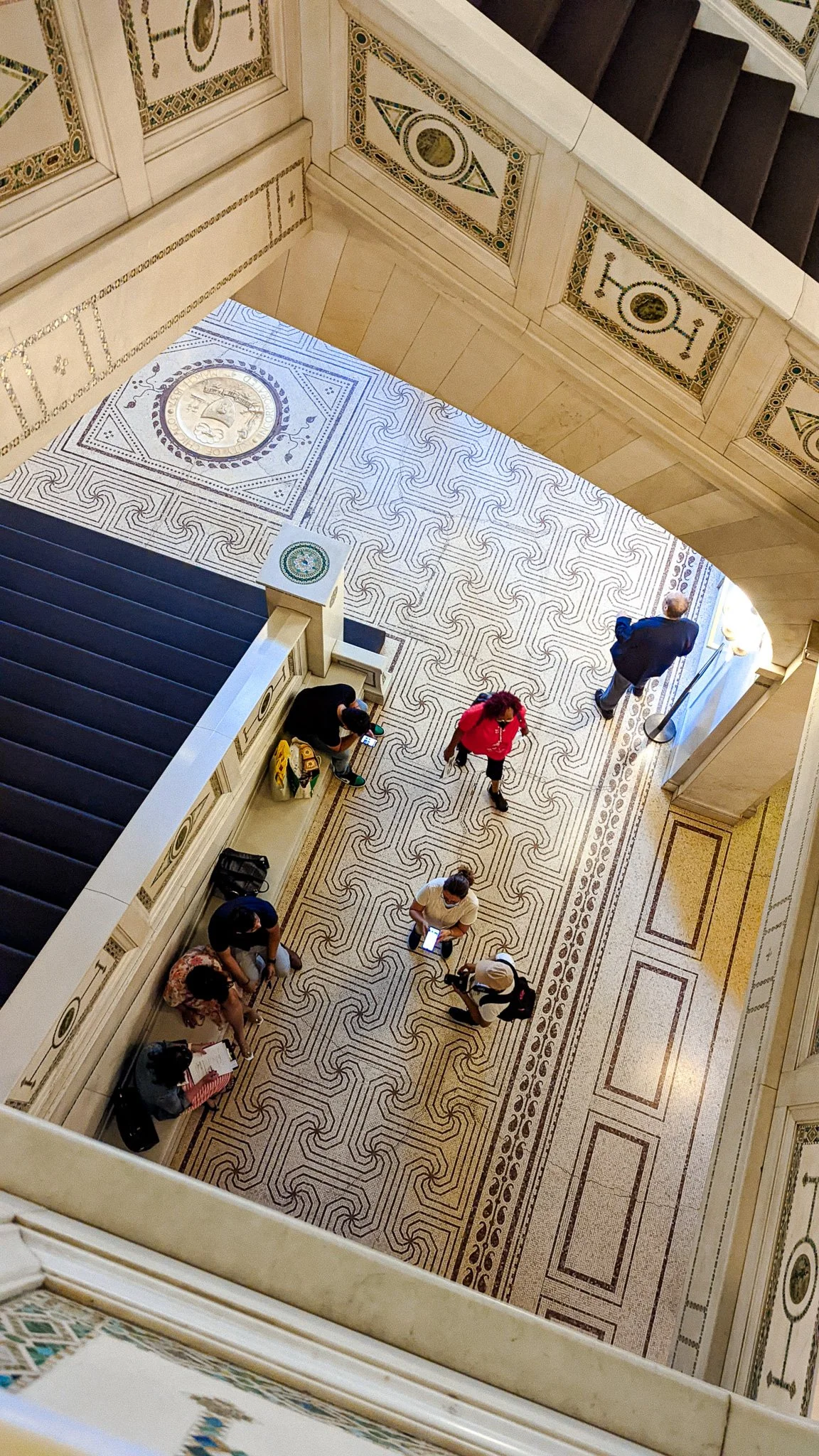 Looking down from a staircase, people walking on a decorative tile floor in a building with ornate wall and ceiling details.