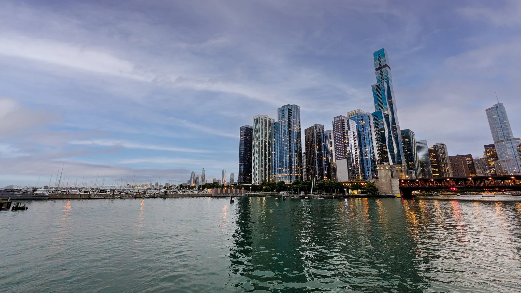 Skyline of a modern city with tall skyscrapers viewed from across a body of water, with boats docked along the shore.