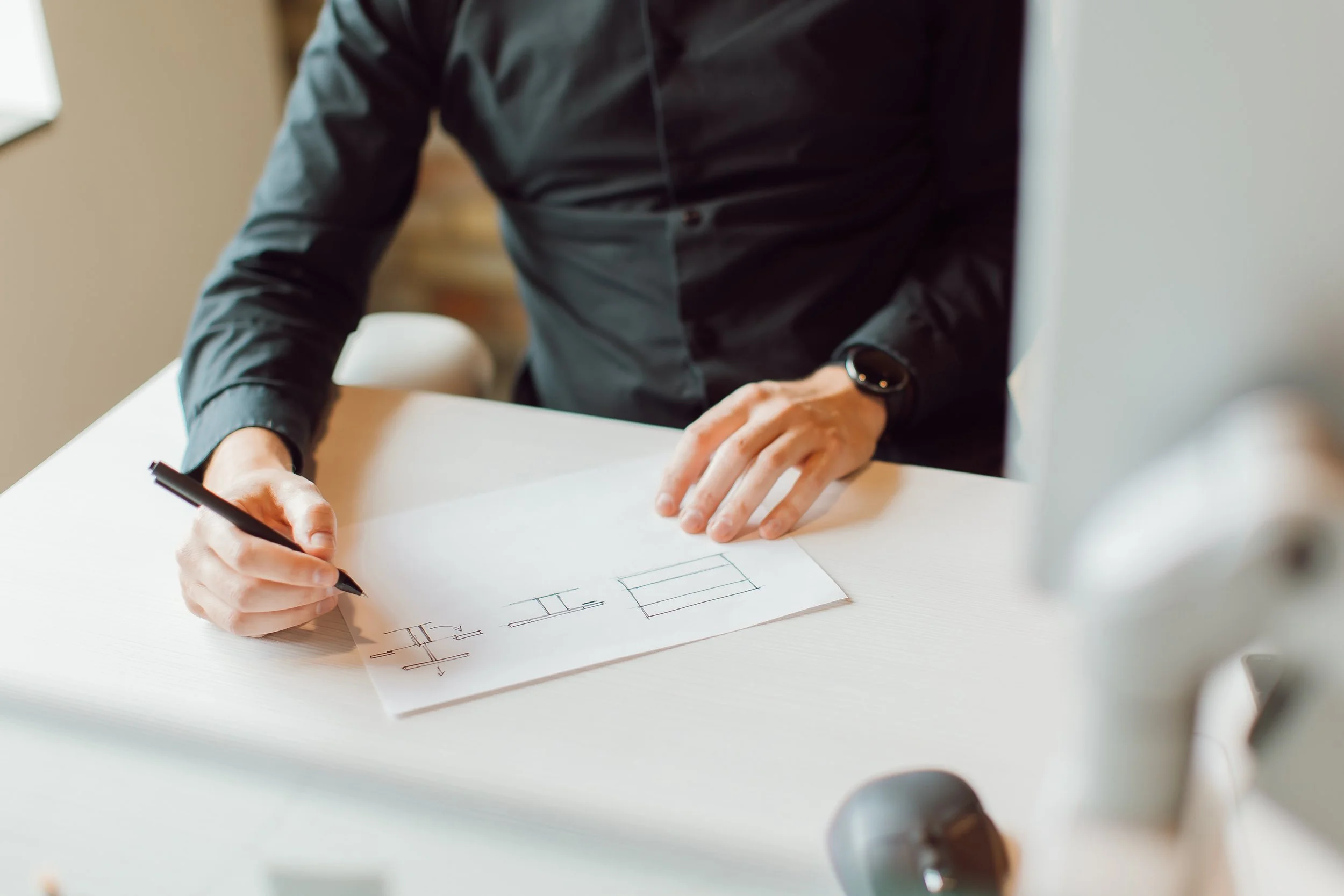 A person in a black shirt is sitting at a table, drawing sketches or designs of furniture on a piece of paper with a pen. The person's left hand rests on the paper, and the right hand holds the pen. The person is also wearing a watch on their left wrist.