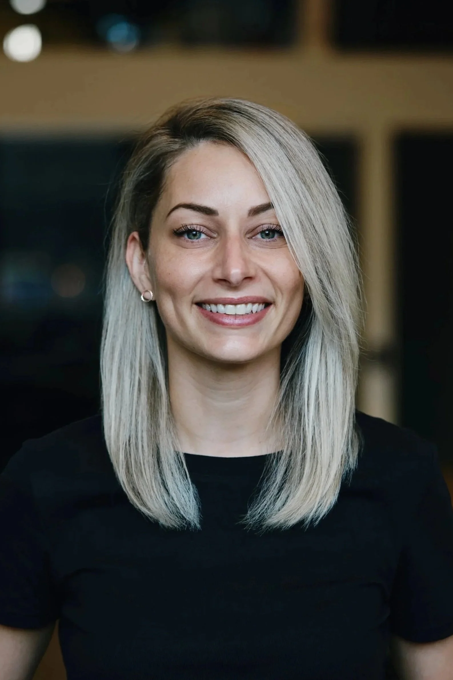 Une femme blonde souriante portant un t-shirt noir, vue de face, en intérieur.
