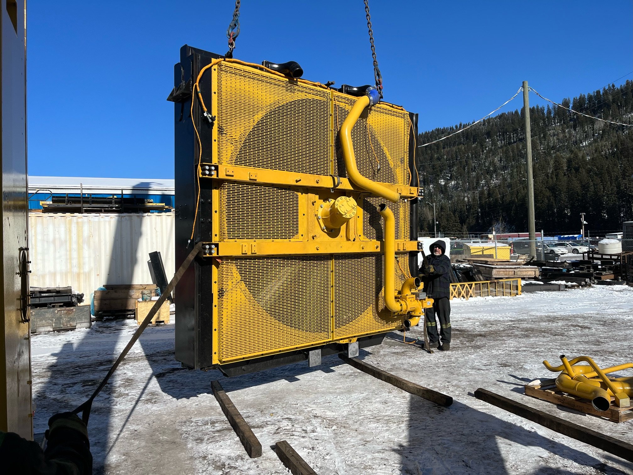 A large yellow industrial radiator or heat exchanger with pipes, supported by a metal frame, outdoors in a snowy area. A person in dark work clothing and safety glasses stands beside it, holding a hose. In the background, there are storage containers, pallets, vehicles, and snow-covered trees under a clear blue sky.