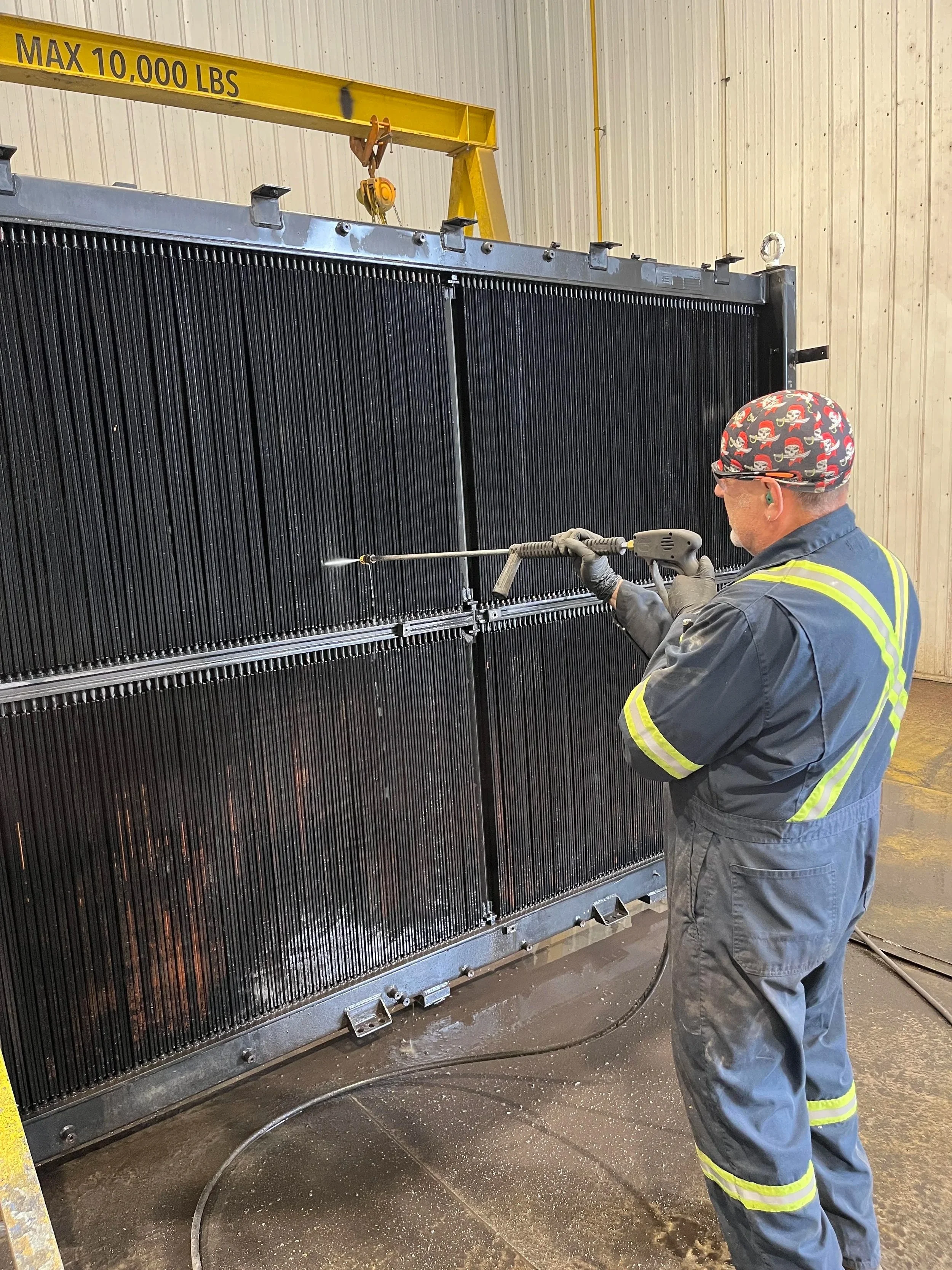 A worker in a safety uniform and a bandana cleaning a large black metal radiator with a high-pressure water hose inside an industrial building.