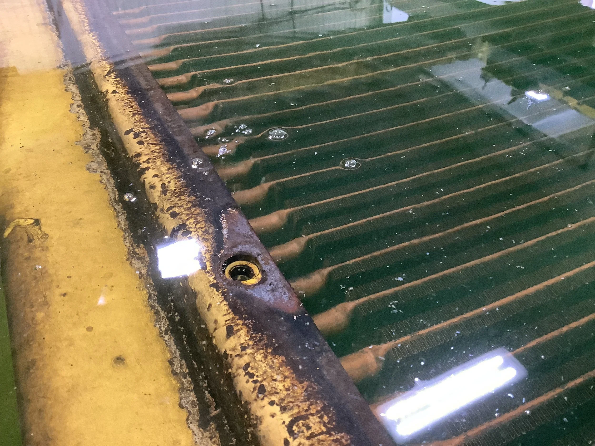 A close-up of a rusty metal grate on a yellow surface, with water and bubbles on the grate.