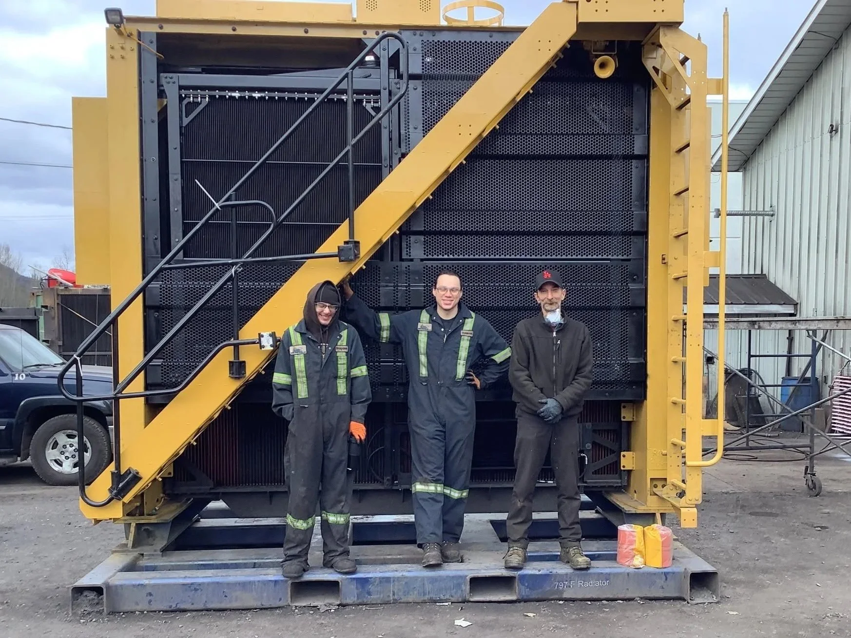 Three workers standing in front of large industrial equipment outdoors, with vehicles and a building visible in the background. The workers are dressed in work uniforms and safety gear.