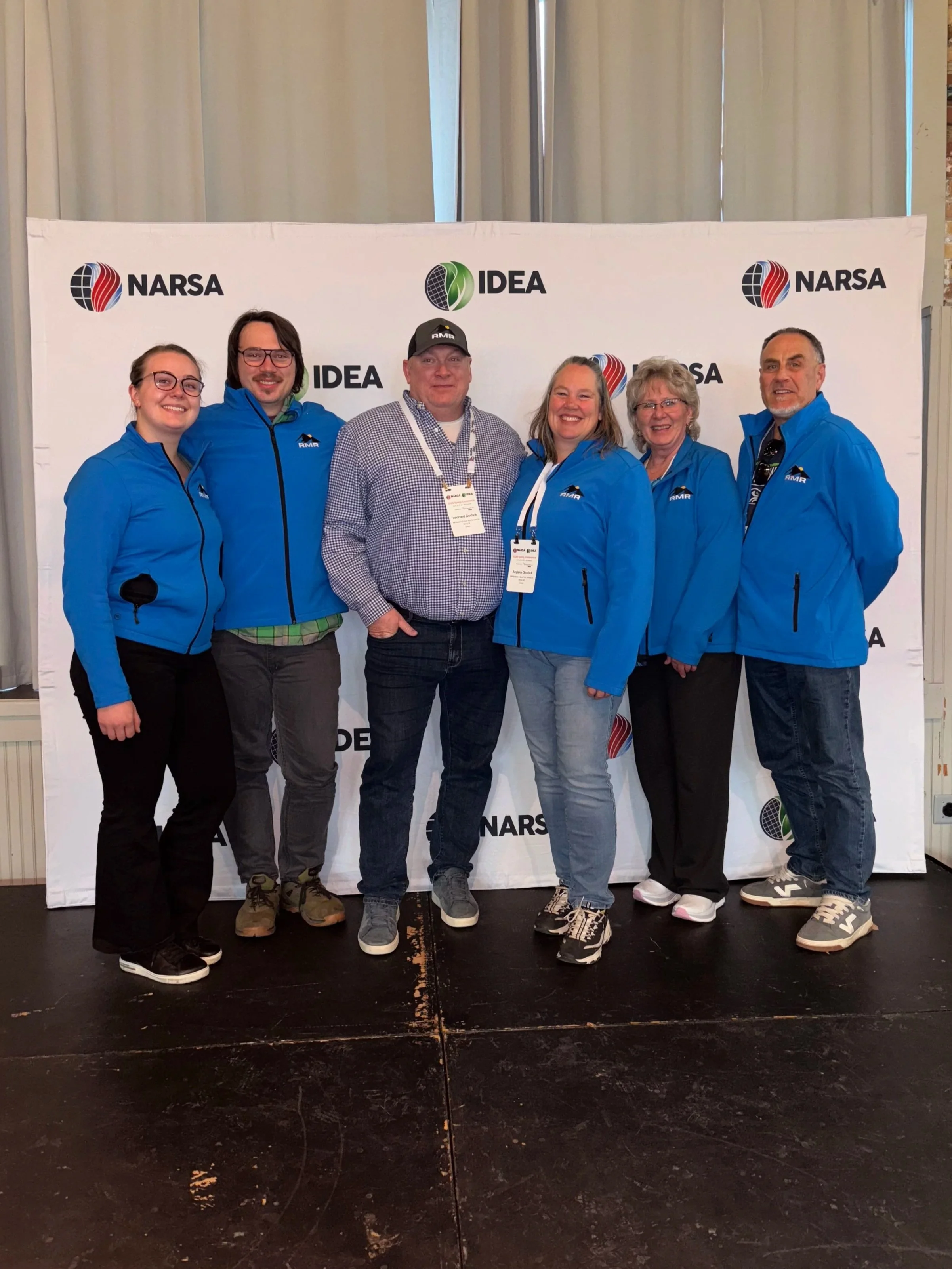 Group of six people dressed in blue jackets standing in front of a white backdrop with logos for NARSA and IDEA at an indoor event.