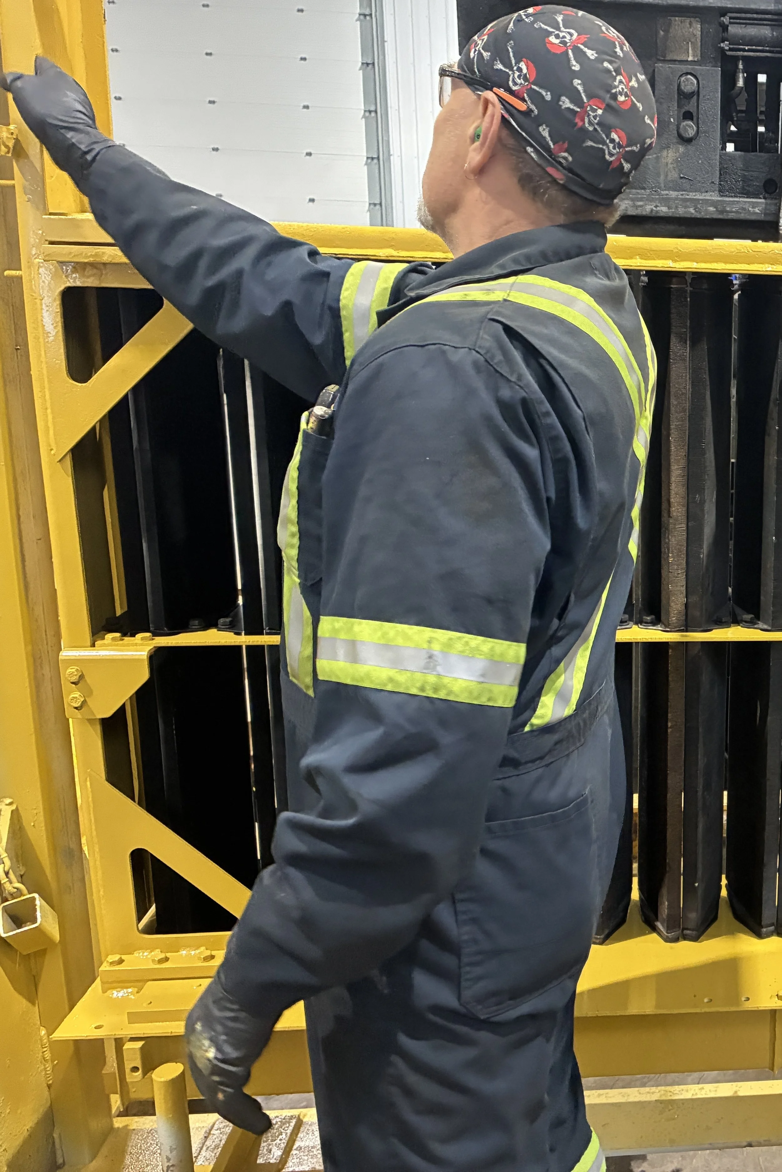 A worker in safety gear, including gloves, a high-visibility vest, and a skull-patterned cap, is inspecting or operating machinery in an industrial or manufacturing setting.
