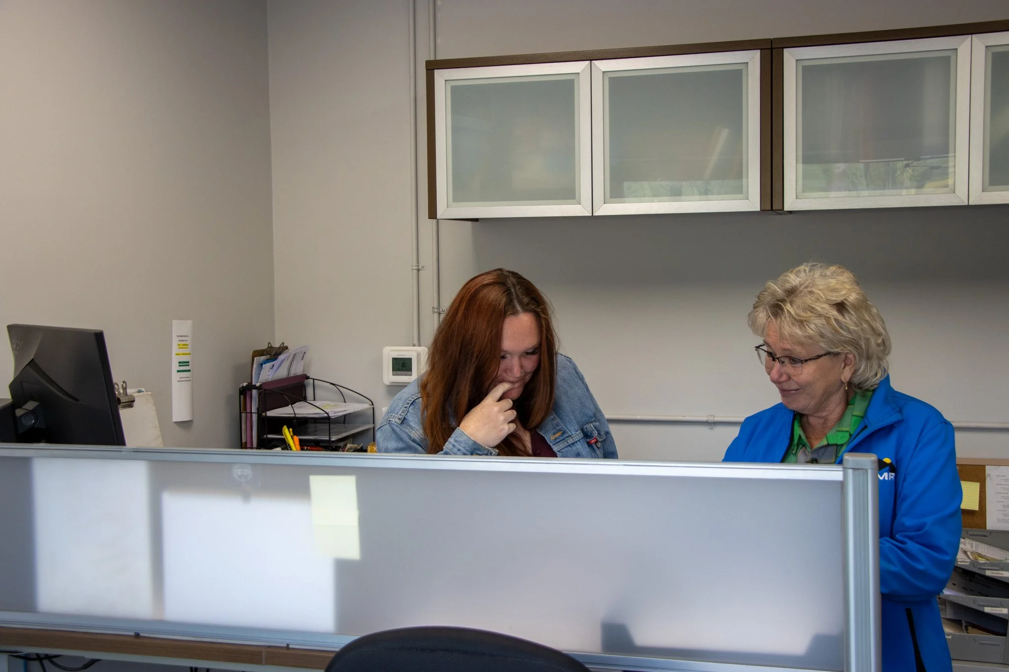 Two women sitting at a desk in an office, engaged in conversation. One has reddish hair and is wearing a denim jacket, the other has gray hair and glasses, wearing a blue shirt. Office supplies and a computer monitor are on the desk, with cabinets on the wall behind them.