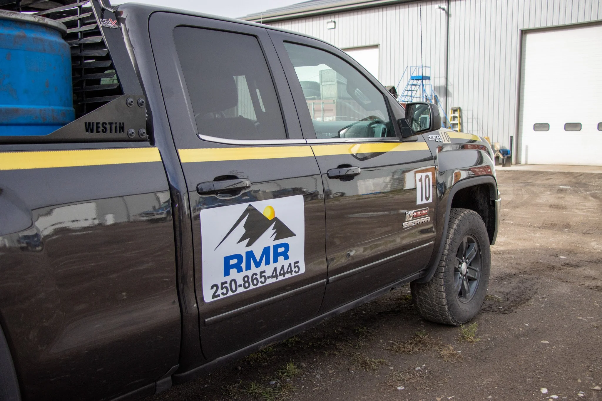 A black pickup truck with a yellow stripe parked outside a large industrial building with a white roll-up door. The truck has a logo and phone number on the door, along with tools and equipment in the truck bed.
