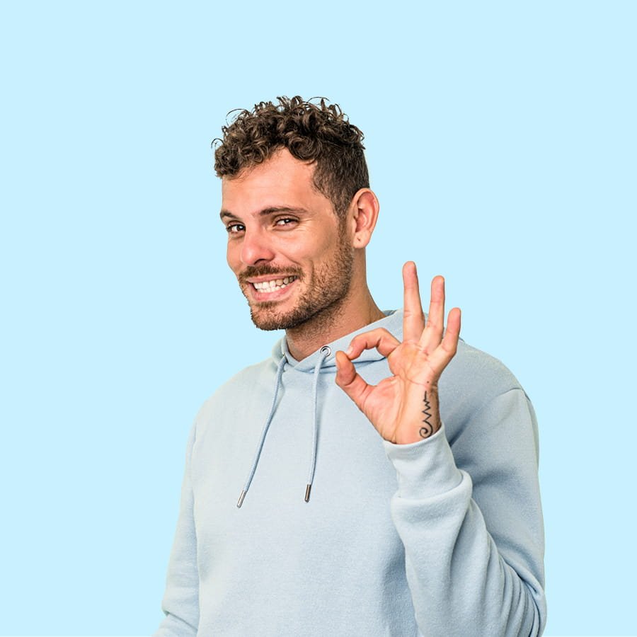 A man with curly brown hair and a beard making an OK hand gesture and smiling, wearing a light blue hoodie against a light blue background.