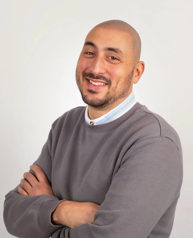 A smiling man with a beard and short hair, wearing a gray sweater and a collared shirt, standing with arms crossed against a plain background.