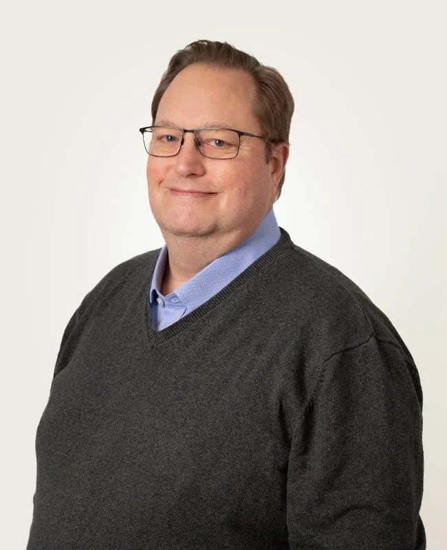 Portrait of man with glasses, wearing a blue collared shirt and a dark grey sweater, smiling at the camera in a studio against a plain background.