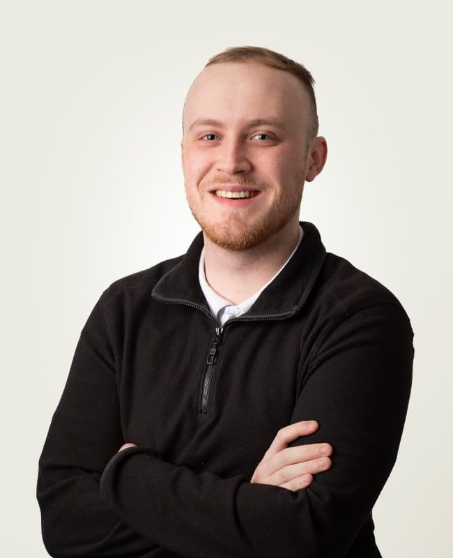 Portrait of with short blond hair and a beard, wearing a black zip-up jacket over a white shirt, smiling with arms crossed against a plain background.