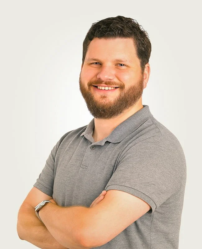 Portrait of a man with brown hair and beard smiling, wearing a gray polo shirt with arms crossed against a plain background.