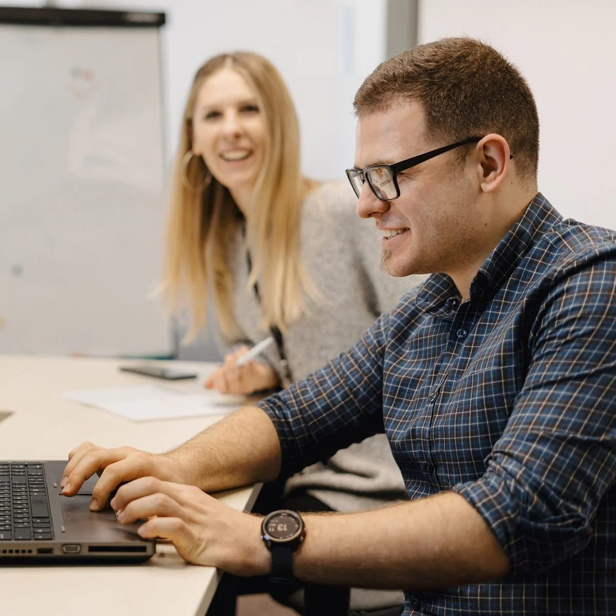 A man wearing glasses and a plaid shirt working on a laptop, smiling, while a woman with long blonde hair and hoop earrings looks on and laughs in the background.