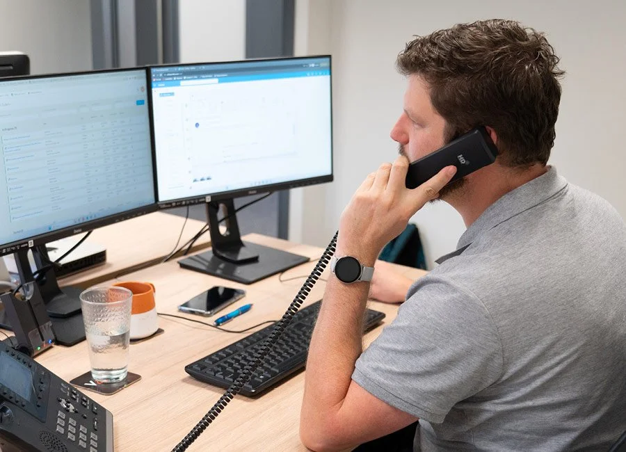 Man on the phone at a desk with dual monitors, a glass of water, a phone, a keyboard, and various office supplies.