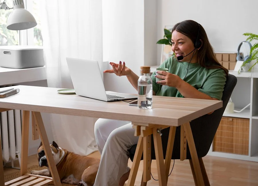 A woman with a headset sitting at a wooden desk, smiling and gesturing during a video call, with a small dog lying underneath the desk.