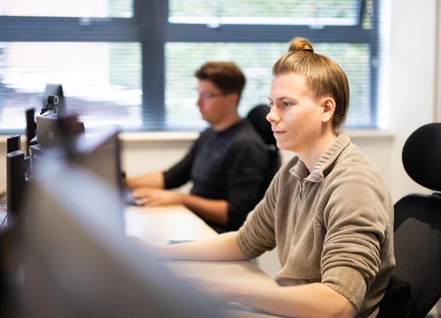 A woman with a ponytail working at a computer in an office with other employees in the background.
