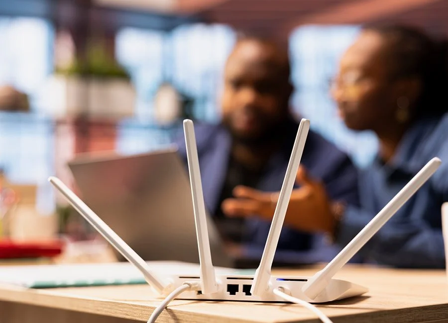 Wireless router with four antennas on a desk in an office, with two blurred people working in the background.