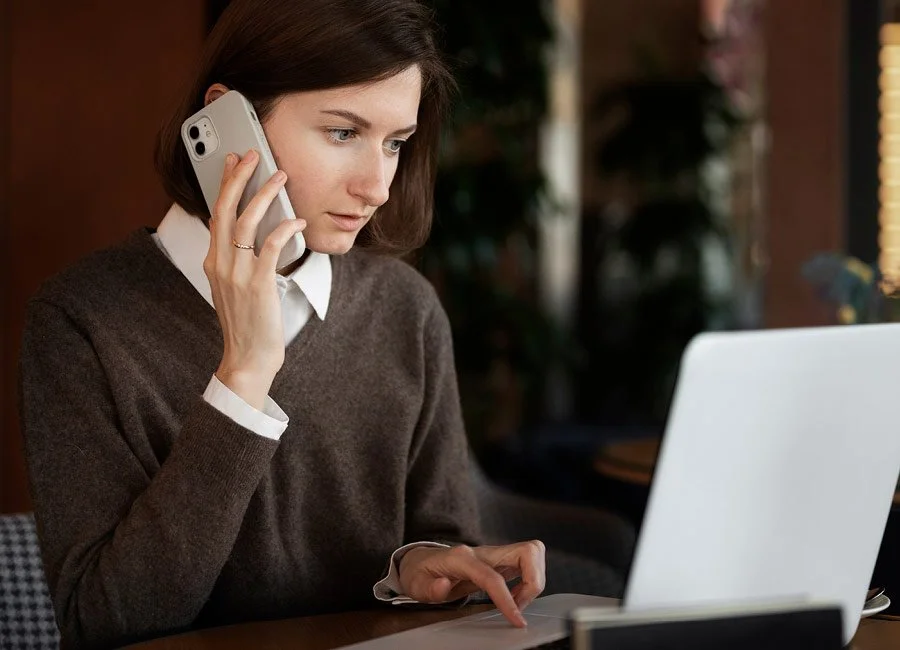 A woman with dark hair wearing a brown sweater and white collared shirt talking on a smartphone while working on a laptop in a cozy indoor setting.