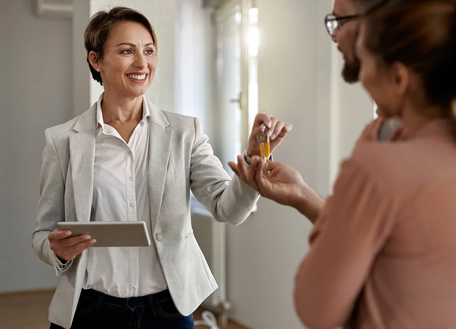 A woman in a light gray blazer hands over house keys to another woman, who is smiling, in a bright residential setting.
