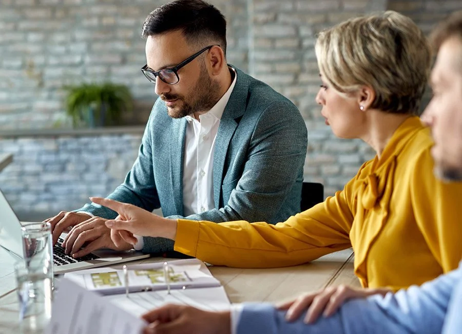Three business professionals in discussion at a meeting table, with documents and a laptop in an office setting.