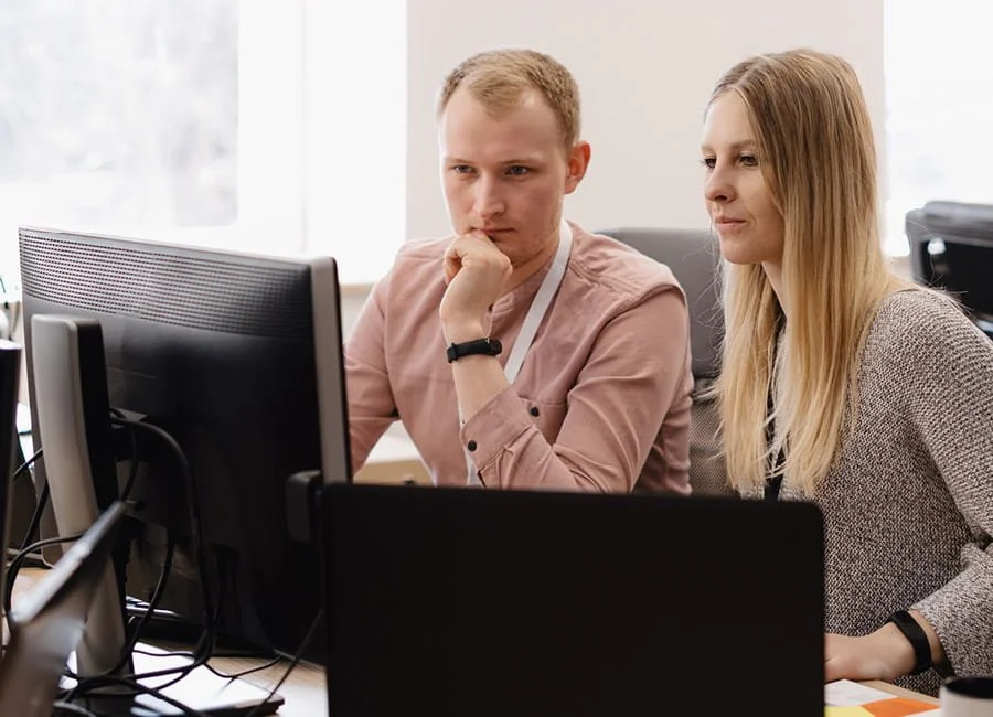 Two coworkers, a man and a woman, working at a desk with computers in an office.