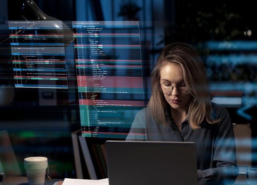 Young woman wearing glasses working on a laptop in a dark room with code and data on transparent screens in the background.