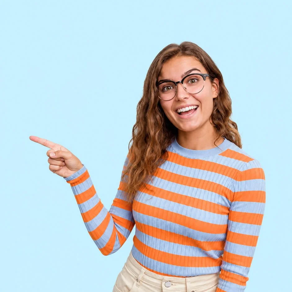 A young woman with curly brown hair wearing glasses, a striped orange and blue sweater, and beige pants, smiling and pointing to her left against a light blue background.