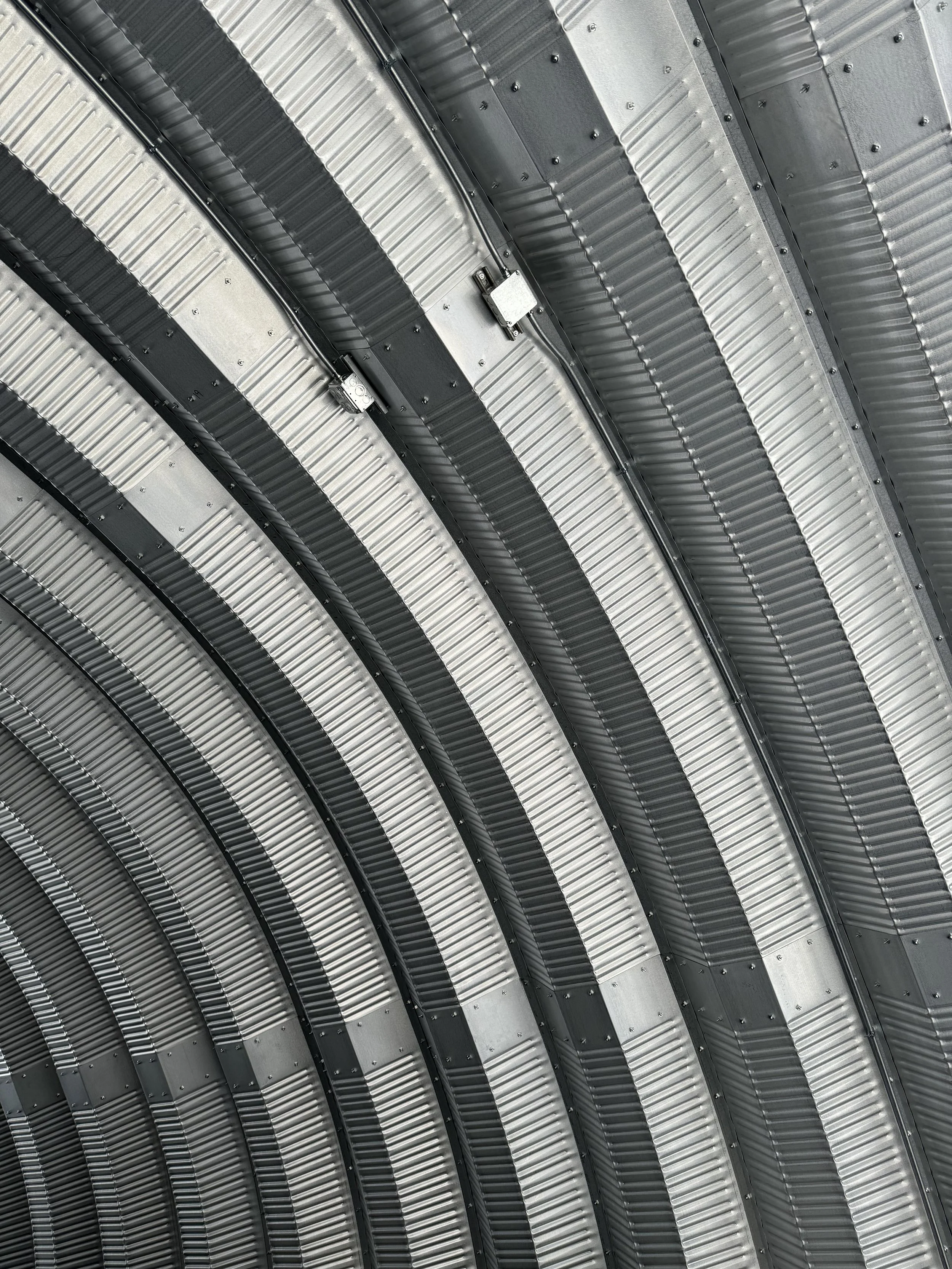 Close-up of a curved metallic roof structure with parallel ridges and several electrical or lighting fixtures attached.