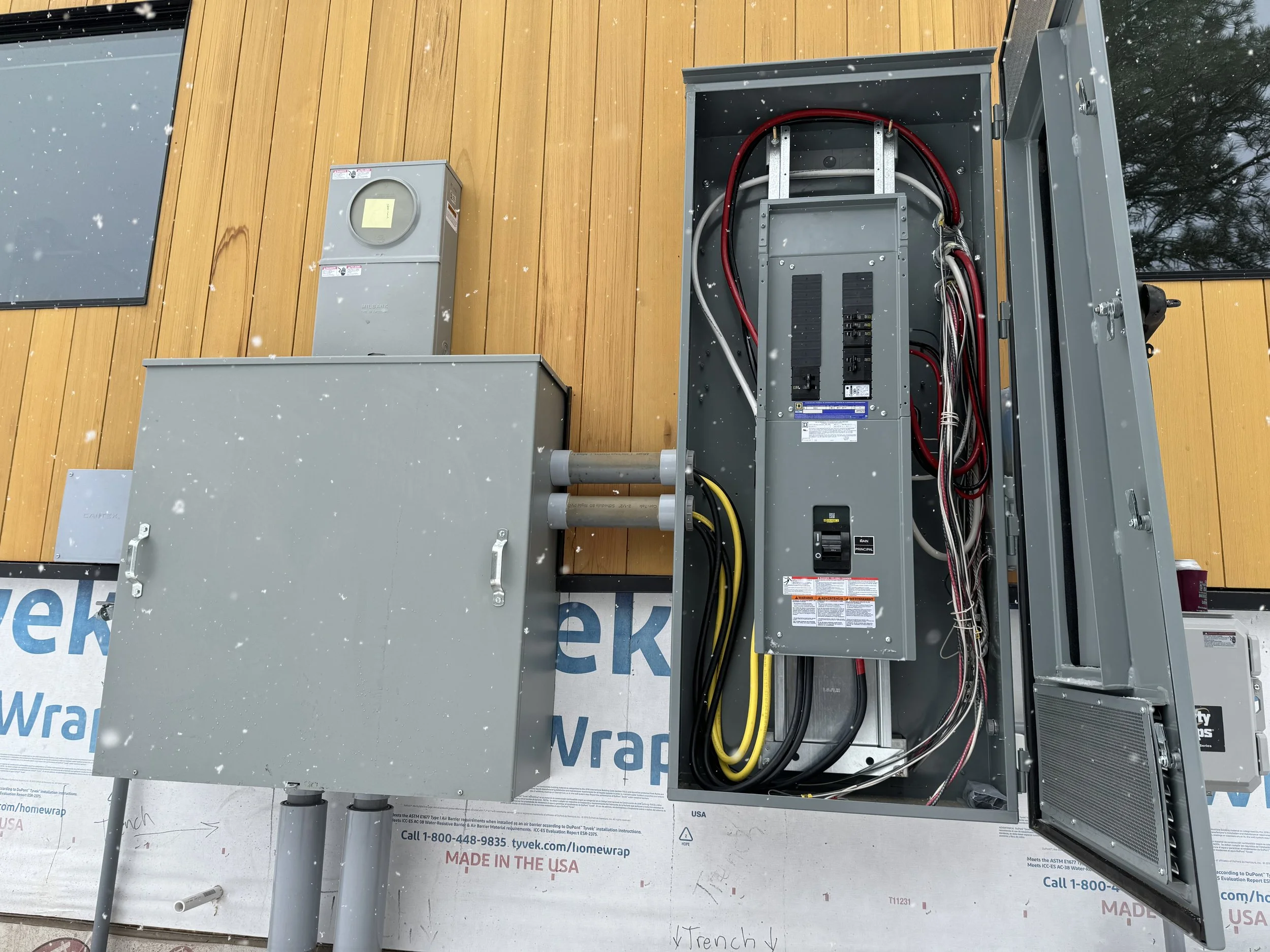 Electrical meter and utility box installed on a wooden wall with an open electrical panel showing wiring.