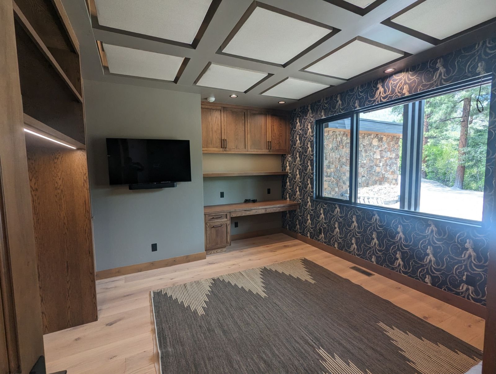 Empty room with built-in wooden desk and cabinets, large window with patterned curtains, patterned wallpaper, ceiling with recessed lights, hardwood floors, and a flat-screen TV mounted on the wall.
