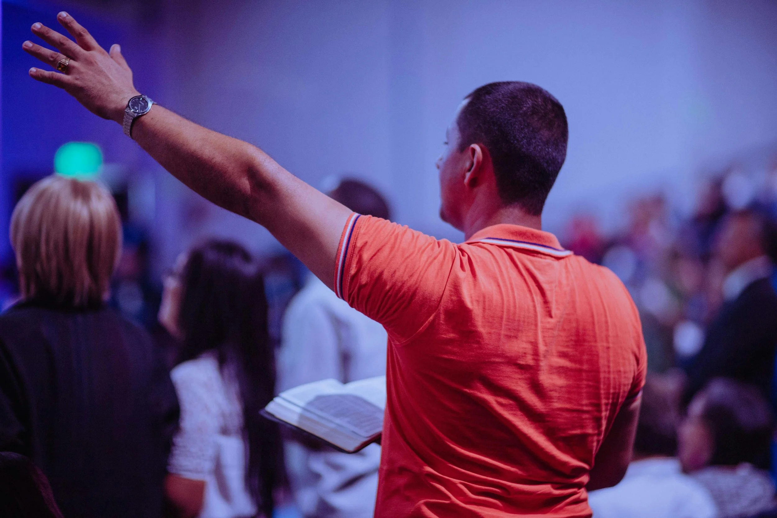 A person in an orange shirt with a watch on their left wrist is raising their hand during an event or conference. The background shows other attendees, with some looking at a speaker or presentation, in a dimly lit room with purple lighting.